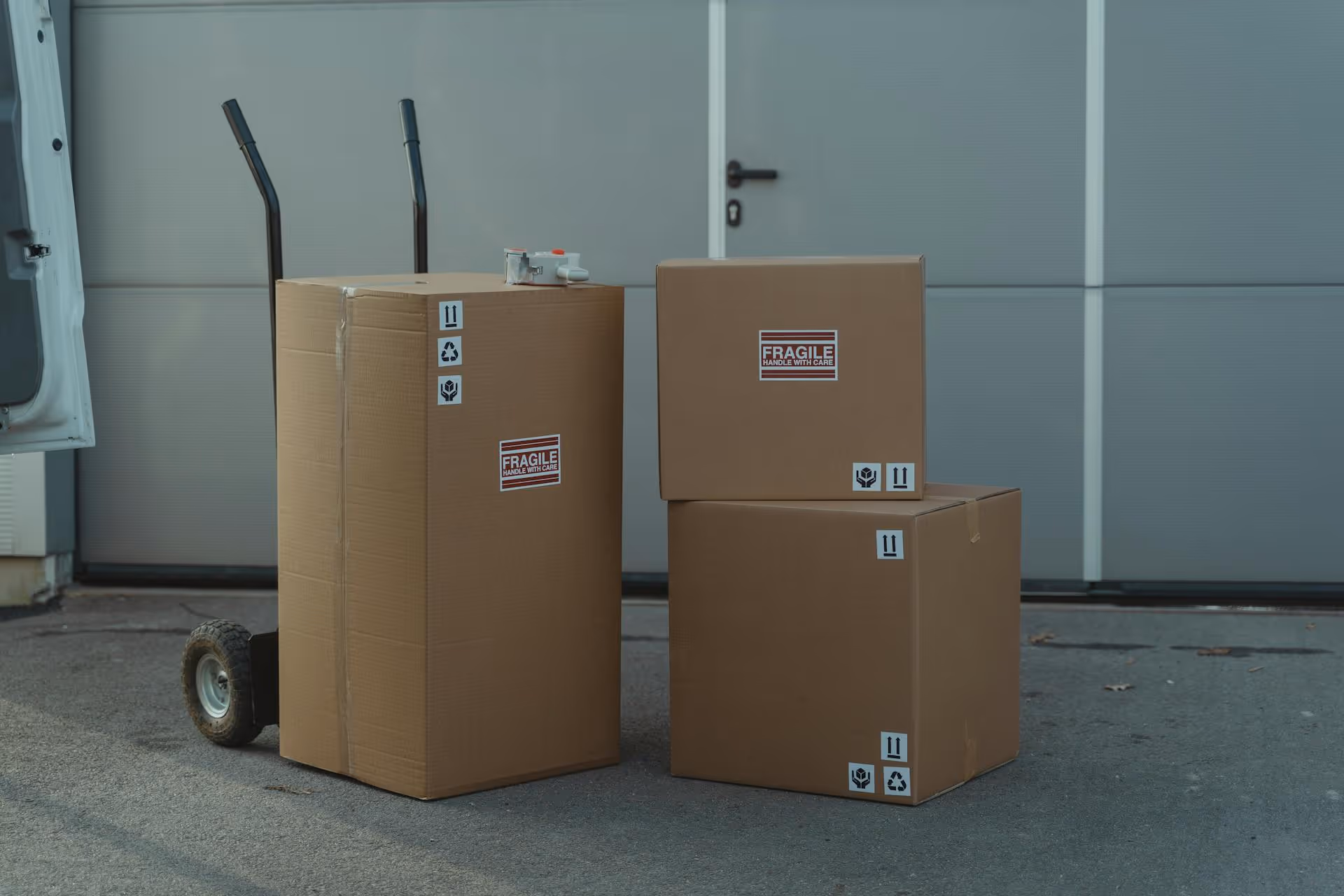 Three brown cardboard boxes labeled fragile and with handling icons, two stacked and one on a hand truck near a closed door.
