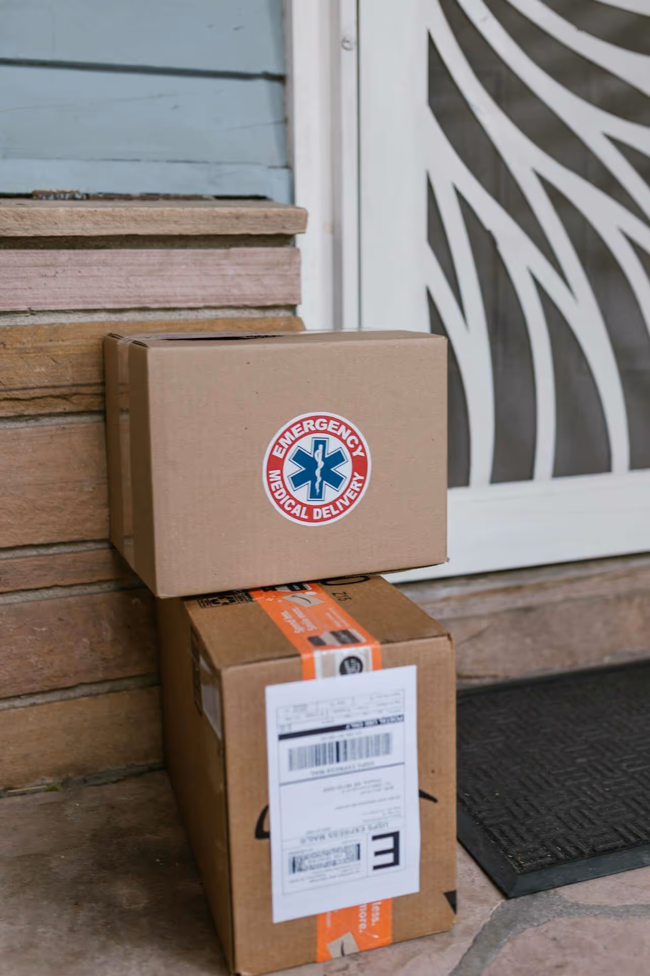 Two cardboard boxes on a doorstep, the top one labeled Emergency Medical Delivery with a blue star of life symbol.