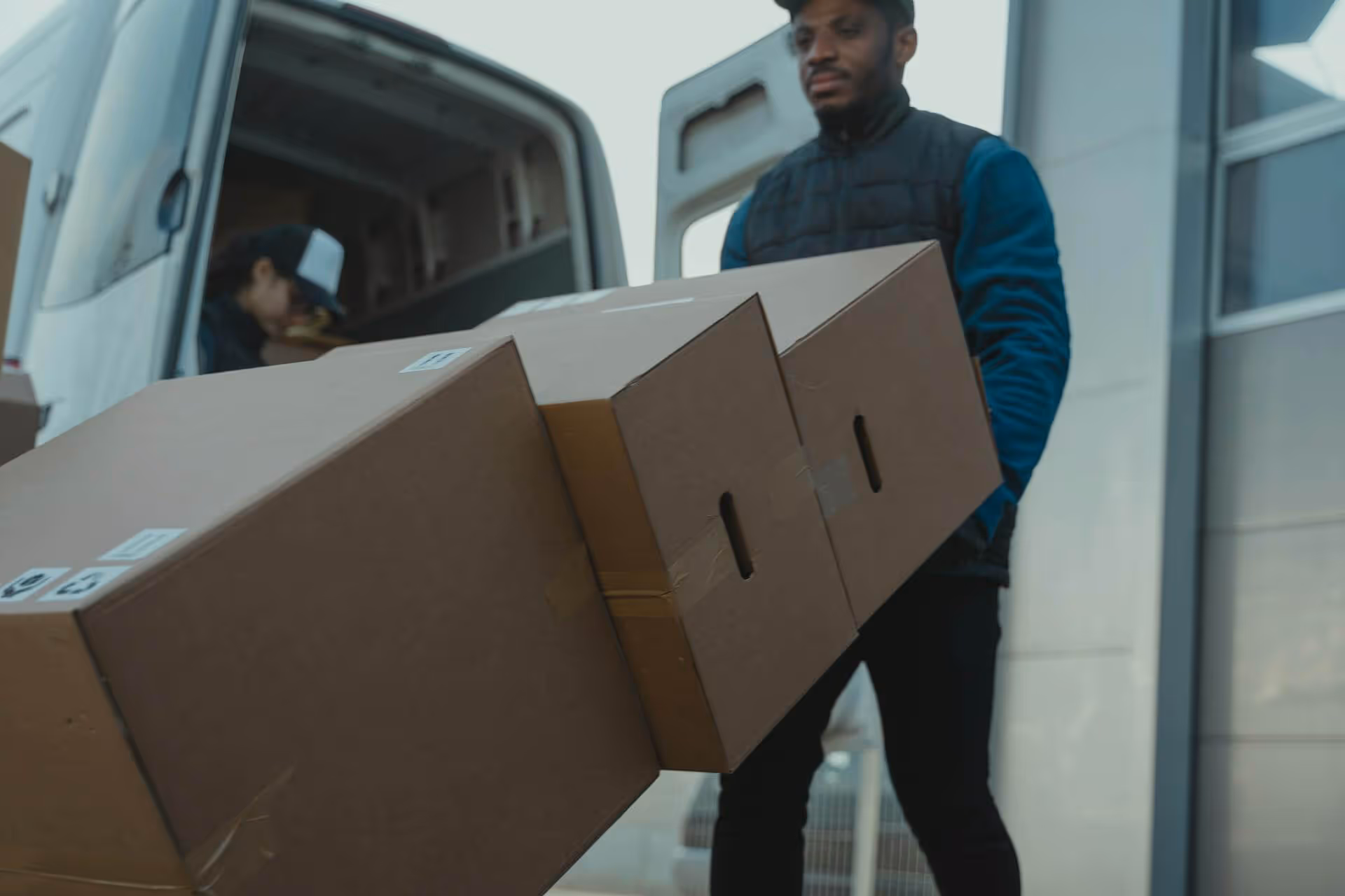 Delivery worker carrying large cardboard boxes outside a delivery van with another person in the background.