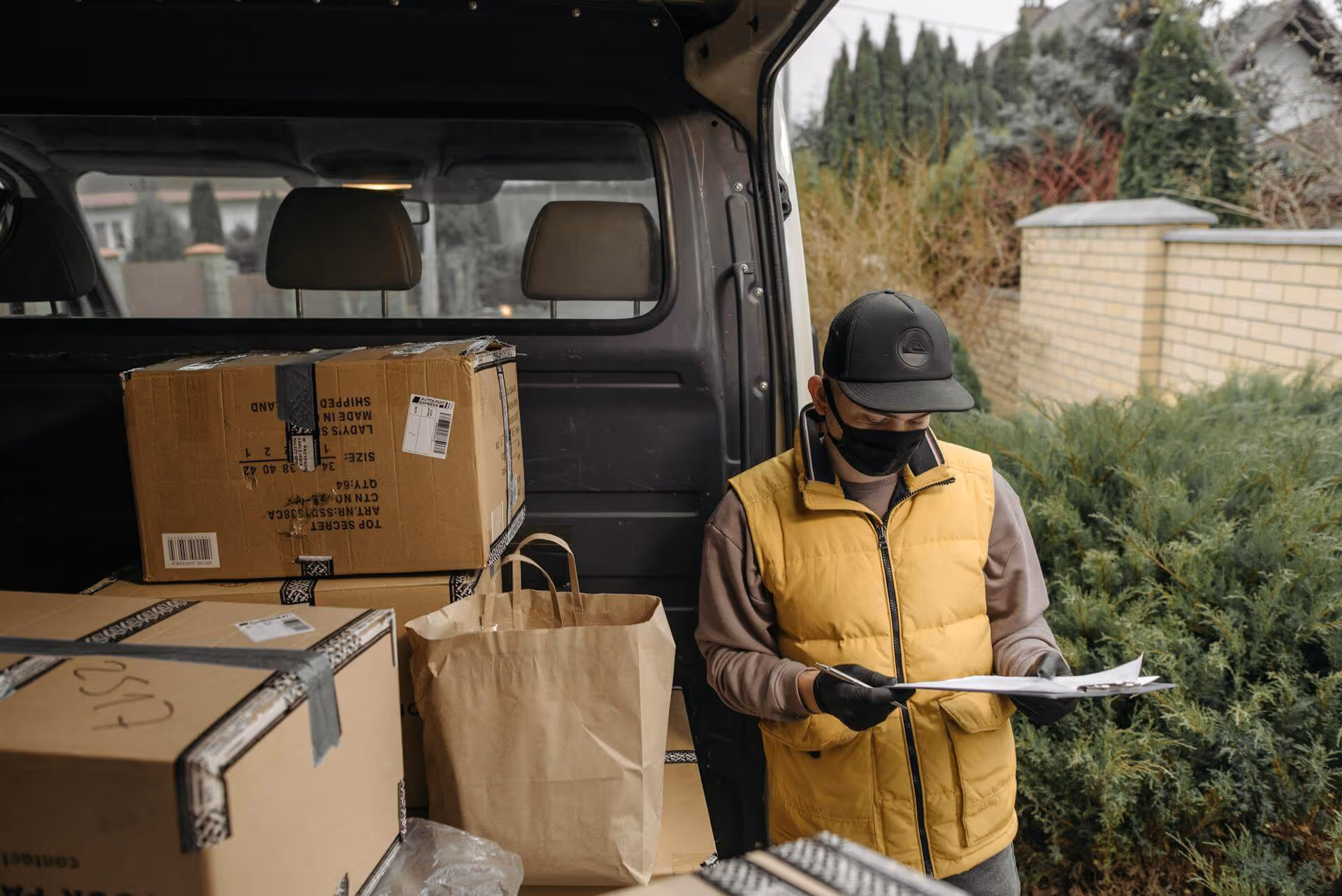 Delivery person wearing a yellow vest and black face mask checking a clipboard near the open back of a van loaded with cardboard boxes and a paper bag.