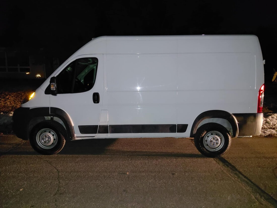 White cargo van parked on a street at night, visible from the side with one headlight on.