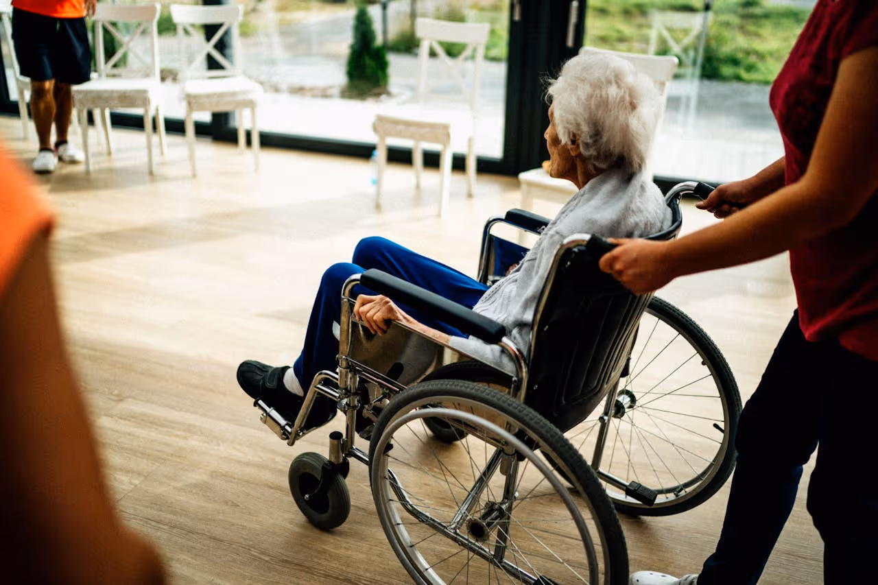 A person in blue pants and a gray sweater is seated in a wheelchair being pushed by another individual indoors.
